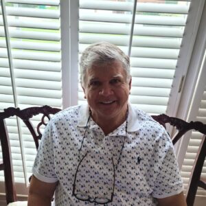 Smiling elderly man sitting in a chair by a window with blinds.